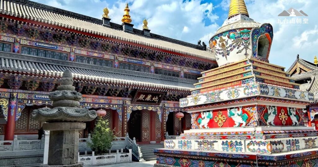 這張照片拍攝於五台山的殊像寺,展現了濃厚的藏傳佛教特色。圖片中央是一座華麗的白塔,塔身裝飾著精緻的彩繪圖案,包括獅子、蓮花與吉祥紋飾,象徵佛法的吉祥與莊嚴。背景中的大殿雕梁畫棟,色彩鮮豔,充滿藏傳藝術的細膩風格,屋頂上的金色裝飾更增添了一份神聖感。整體氛圍莊嚴而華美,是五台山藏傳佛教信仰的重要場域。