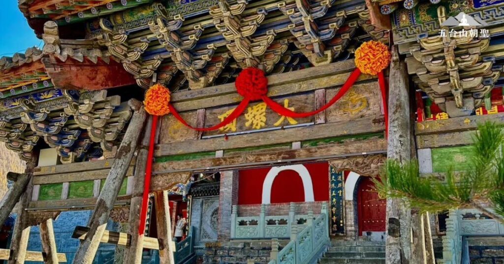 這張圖片展示了五台山大螺頂寺（Dailuo Temple）的山門，木製牌匾上書「大螺頂」，象徵其特殊的地理位置與宗教意義。門楣上懸掛著紅色與橙色的吉祥裝飾，增添了莊重與喜慶的氛圍。大螺頂寺位於五台山頂部，是朝聖者必訪的重要寺廟之一，信徒需攀登長階而上，象徵修行路上的堅持與突破。