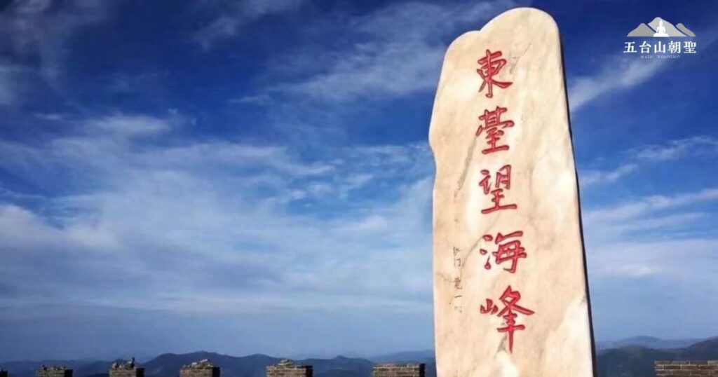 東台望海峰是五台山五大台之一，以其壯觀的日出景色聞名。照片中，一塊巨大石碑矗立於高山之巔，上刻「東台望海峰」紅色大字，顯得格外醒目。藍天白雲映襯下，遠處山巒連綿起伏，視野遼闊，讓人感受到登頂後的寧靜與遼闊天地的震撼。此處是朝聖者和遊客的熱門打卡點，也被譽為觀賞佛光奇景的理想地點。