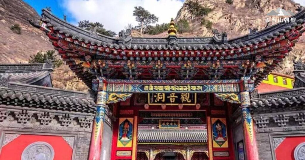 觀音洞位於五台山，寺院入口以華麗的紅色、金色裝飾，搭配精緻的雕刻與彩繪，展現濃厚的藏傳佛教風格。門額上書「觀音洞」，象徵這座寺廟與觀世音菩薩的深厚淵源。建築外圍環繞著堅固的灰磚牆，牆面上的圓形雕刻突顯佛教文化的細膩藝術。背景是陡峭的山壁，讓整座寺廟彷彿隱身於大自然中，為信徒提供一處靜謐的修行與祈願之地。