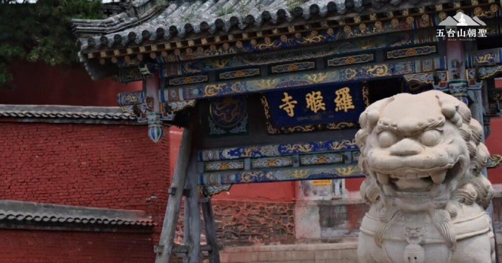 這張圖片展示了五台山羅睺寺（Luo Hou Temple）的精美大門，門額上書寫「羅睺寺」，其建築細節展現出中國傳統佛教寺院的藝術風格。門前的石獅威嚴矗立，象徵守護與吉祥，紅磚牆與藍色牌匾形成鮮明對比，使整體氛圍莊嚴肅穆。羅睺寺是五台山的一座歷史悠久的佛教寺廟，深受信徒與遊客的敬仰與參訪。