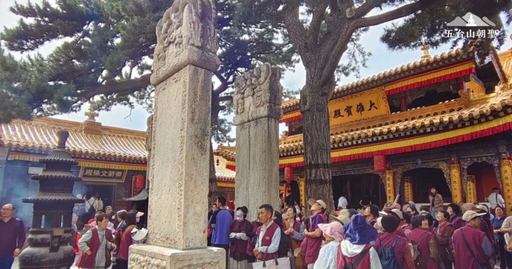 這張圖片捕捉了五台山某座著名寺廟的熱鬧朝聖場景，信徒們穿著統一的朝聖服飾，聚集在寺院前虔誠祈禱。畫面中，兩根雕刻精美的石碑矗立在松樹下，見證著歲月的洗禮，寺廟建築金碧輝煌，屋簷上懸掛著紅黃相間的裝飾，增添莊嚴氛圍。背景的香煙裊裊，展現佛教聖地的虔敬氛圍。這張圖片完美呈現五台山作為佛教名山的莊嚴與神聖，吸引來自世界各地的信眾前來朝拜與參訪。