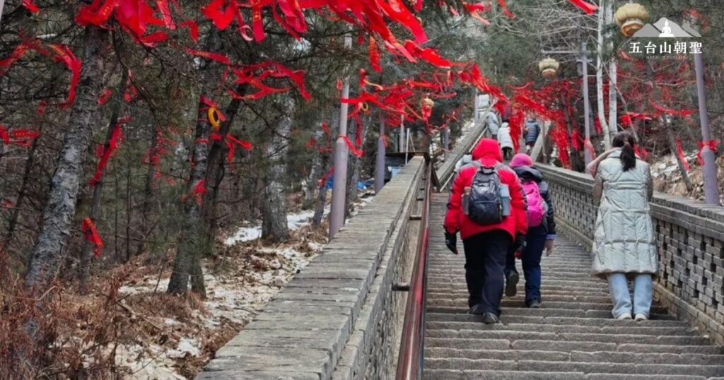 這張照片展現了五台山朝聖者踏上的信仰之路。登山階梯兩側掛滿了祈福的紅色絲帶,在風中飄揚,象徵著信眾的願望與祝福。信徒們身穿厚重衣物,沿著台階緩步而上,展現出對朝聖之旅的虔誠與堅持。冬日的山林間,積雪點綴,為這條靜謐而莊嚴的道路增添了一抹詩意。這條通往靈山的階梯,不僅是一段身體的攀登,更是一場心靈的洗禮。