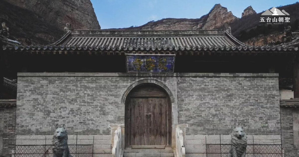這張圖片展示了五台山秘密寺的古樸大門，寺廟位於山谷之中，四周環繞著險峻的岩壁，顯得格外幽靜神秘。寺門以青灰色磚牆砌成，門額上掛有「祝聖寺」的匾額，木質大門緊閉，增添了一絲莊嚴與神秘感。門前兩側擺放著石獅雕像，象徵守護寺院的神聖力量。這座寺廟低調且極具歷史韻味，為五台山的隱秘佛教聖地之一，吸引尋求內心平靜與修行的信眾前來朝聖。