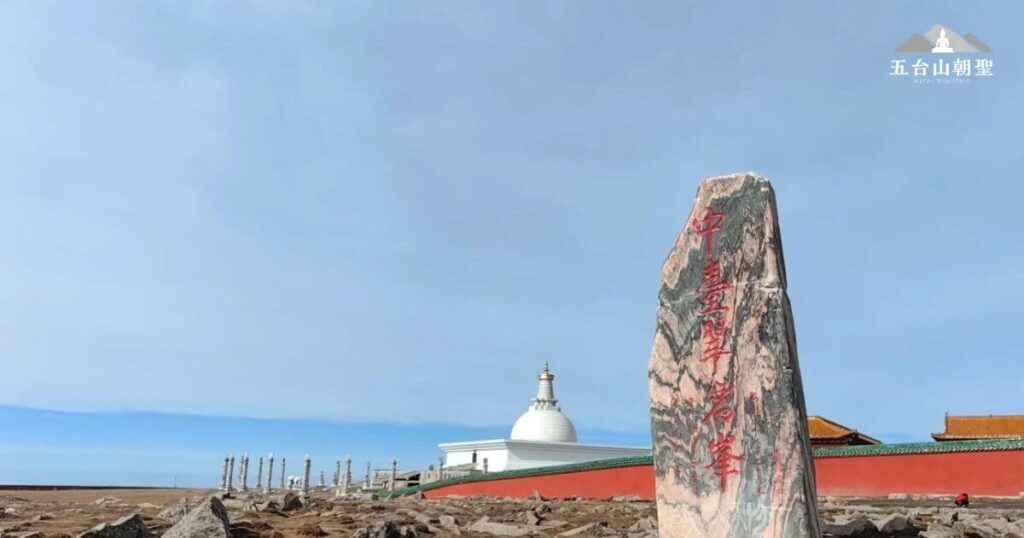 這張照片展示了五台山五個主要高峰之一——中台（翠巖峰）的標誌性石碑，石碑上刻有「中台翠岩峰」幾個紅色大字，顯示出其佛教聖地的地位。在石碑後方，可以看到白塔與寺廟建築，象徵著這座聖山的宗教與歷史價值。周圍地勢開闊，散落的岩石與藍天形成鮮明對比，使整個場景更顯莊嚴與靜謐。這裡是許多朝聖者與登山者尋求智慧與平靜的地方。