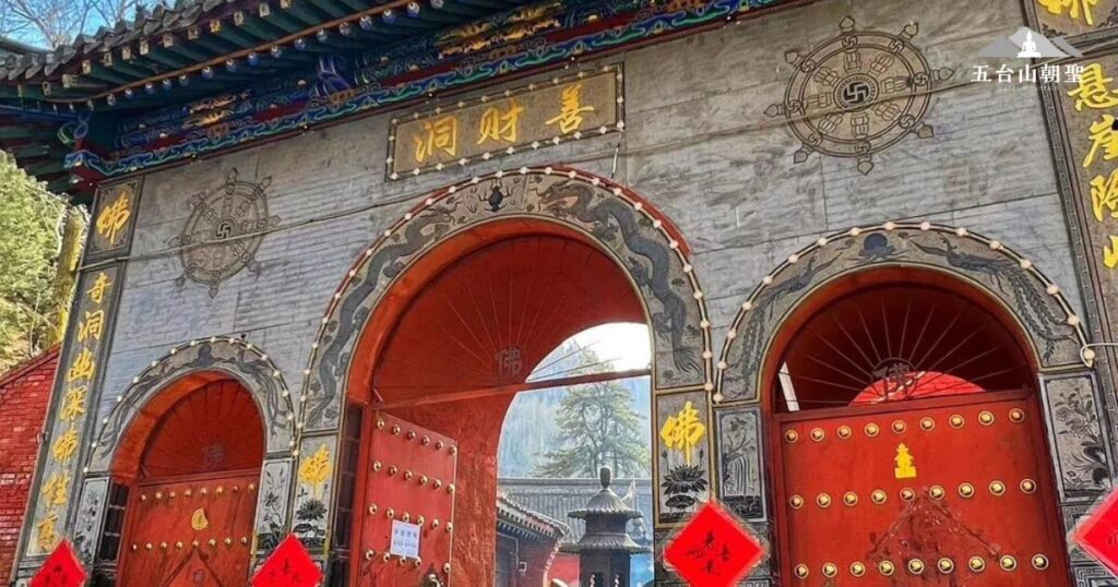 這張照片展示了五台山著名的「聚財洞」入口，其建築風格融合了傳統佛教寺廟的精美雕刻與對稱設計。紅色大門鑲嵌金色門釘，並刻有精緻的圖案，展現莊嚴與莊重。門楣上方刻有「聚財洞」三字，象徵財富與福報匯聚之地。門旁的牆面上裝飾著佛教吉祥圖案，如法輪與佛字，象徵智慧與修行。陽光透過樹梢映照在寺門上，使整體氛圍更顯神聖寧靜，吸引無數信眾前來祈福與參拜。
