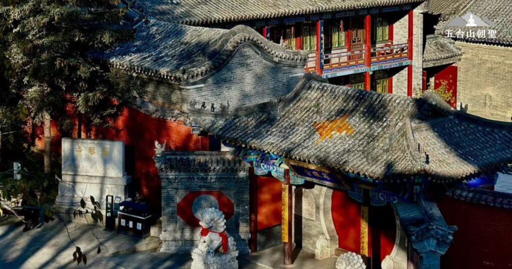 這張照片拍攝的是五台山羅喉寺的門樓，屋簷呈現古樸的灰瓦造型，結構典雅而莊重。門口擺放著一座白色石獅，象徵鎮守寺院的力量，旁邊還有一塊石碑刻有「羅喉寺」字樣。寺院內部建築融合了傳統中式建築風格，門樓上方可見精緻的彩繪裝飾，展現佛教建築的藝術之美。陽光灑落在屋簷之上，使整體畫面更顯寧靜莊嚴，吸引無數信眾前來朝聖與參拜。
