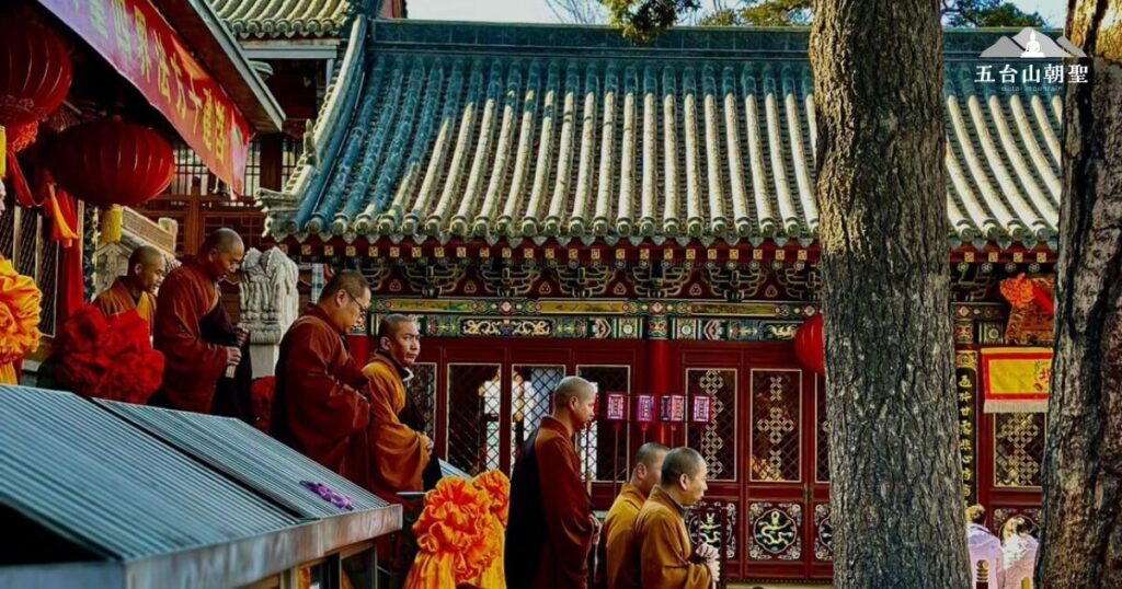 這張圖片展示了五台山（Wutai Mountain）內，數位僧侶身著傳統僧衣，在莊嚴的寺廟建築前進行修行儀式。他們神情莊重，雙手合十，展現出虔誠的佛法信仰。背景是中國古典風格的紅色寺院建築，裝飾著精緻的雕刻與傳統紅燈籠，營造出莊嚴而寧靜的氛圍。五台山作為中國佛教四大名山之一，是許多信眾朝聖與修行的聖地。