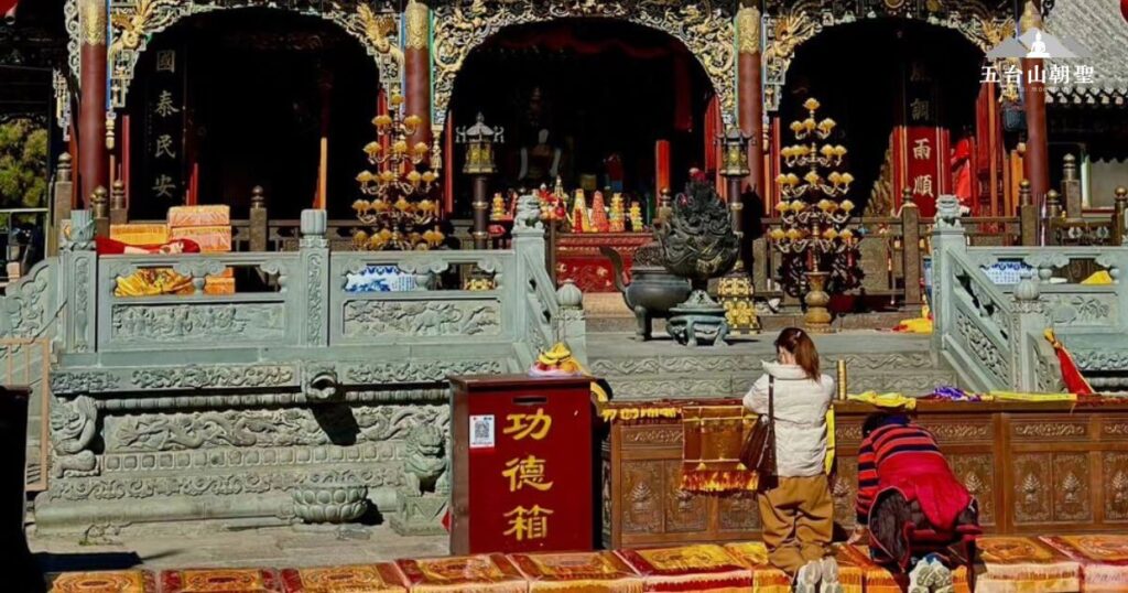 這張圖片捕捉了五台山某座寺廟的朝聖場景,信徒們雙膝跪地,虔誠叩拜於供奉佛像的殿堂前。寺廟外觀華麗,雕刻精美,供桌上擺滿了各式供品與金色燭台,展現莊嚴的宗教氛圍。前方的「功德箱」提醒著信徒行善積福,將善念轉化為實際的修行功德。這裡是五台山信仰與修行的核心,吸引來自世界各地的信眾前來朝拜,尋求內心的平靜與智慧。