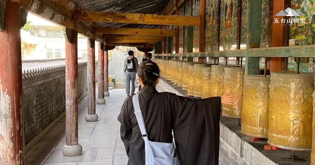 這張圖片拍攝於五台山的一處轉經長廊，信徒沿著長廊緩步前行，手輕觸金色轉經輪，展現佛教傳統的修行方式。長廊內側牆壁上繪有精美的佛教壁畫，描繪佛陀與諸神的故事，為朝聖者帶來視覺與心靈的雙重啟發。木製長廊頂部展現歲月痕跡，見證著歷史的流轉。這裡是信徒感受佛法、沉澱心靈的聖地，每一次轉動經輪，都是對智慧與福報的祈願。