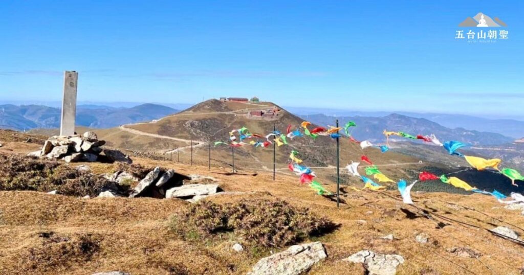 這張照片捕捉到五台山其中一座巔峰的壯麗景色，遠方的寺廟座落於山丘之上，象徵著佛教聖地的寧靜與莊嚴。五彩繽紛的經幡迎風飄揚，傳遞著信徒的祈願，與廣闊的藍天和連綿的山脈相映成趣。這裡不僅是朝聖者心靈寄託之地，也是一個讓人感受天地寧靜、洗滌心靈的所在。