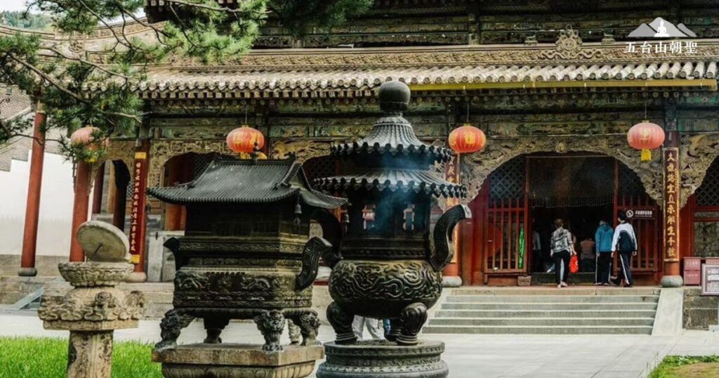 這張照片捕捉了五台山寺廟入口前的香爐，香煙裊裊升起，營造莊嚴肅穆的氛圍。背景中，遊客與信眾正在步入寺廟，紅色燈籠與木雕建築展現中國傳統佛教建築的獨特美學。前方的香爐雕刻精美，象徵佛法的傳承與信仰的虔誠，是五台山朝聖旅程中常見的景象。這座古剎充滿歷史與宗教氛圍，讓每位來訪者都能感受佛教文化的深遠影響。