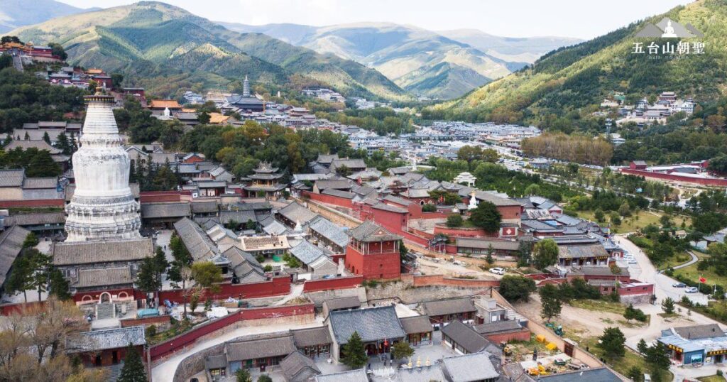 這張圖片展示了五台山（Mount Wutai）的壯麗全景，俯瞰整個山區的寺廟群，層疊的屋頂和遠處的白塔尤為醒目，背景是壯麗的山脈。五台山作為文殊菩薩的聖地，吸引無數信徒前來朝聖。這幅圖片捕捉了五台山的自然與人文景觀，象徵著精神與智慧的追求，讓人感受到一種與天地相連的神聖氛圍，適合用來推廣五台山的宗教與文化魅力。