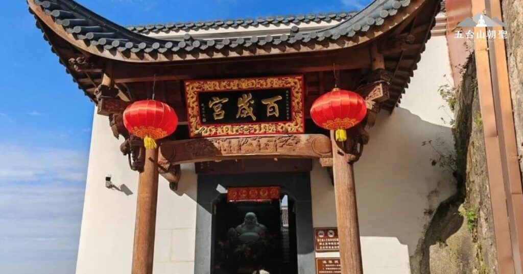 這張照片顯示了一座古老宮殿的門廊，門口懸掛著兩個紅燈籠，增添了傳統的文化氛圍。上方的金色匾額寫著「宮殿百年」，表達了這座宮殿歷史悠久的象徵。門廊結構採用木質材質，古樸而穩重，周圍的細節雕刻精緻，充滿了中國古建築的特色。整個景象呈現出一種莊嚴、神聖的氛圍，吸引人們深入探索其中的歷史與文化。