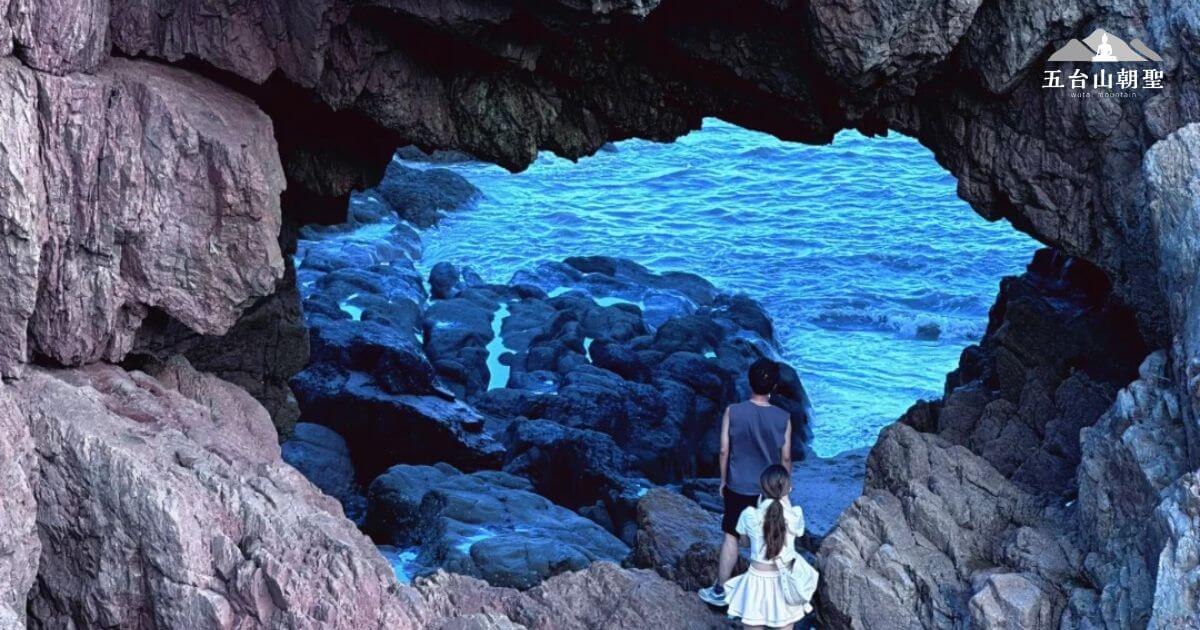 這張圖片呈現了潮音洞（Chaoyin Cave）內部的美麗景觀。一對父女站在洞口，凝視著遠方的海洋，海浪拍打著岩石，背景的海水呈現出神秘的蔚藍色。岩洞的形狀與周圍的岩石形成鮮明對比，突顯出這片自然景觀的壯麗和安靜。