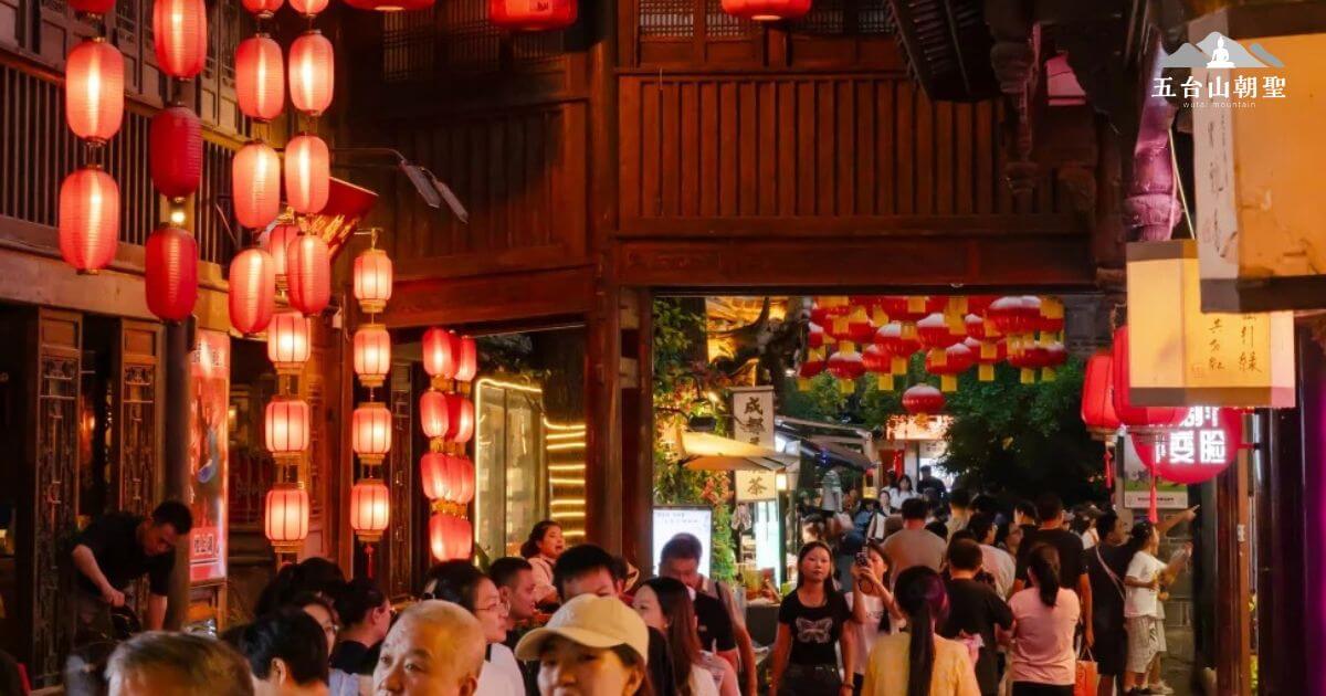 這張圖片展示了成都錦里古街的熱鬧景象。街道兩旁掛滿了紅色燈籠，夜色中燈光映照著來來往往的遊客，氣氛濃厚。周圍的古建築風格保留了傳統的中式特色，攤位上擺滿了各式各樣的小吃和手工藝品，吸引著人們停下腳步細細品味。這是一個融入歷史與現代的完美地點，充滿了人們對成都的熱愛與探險精神。