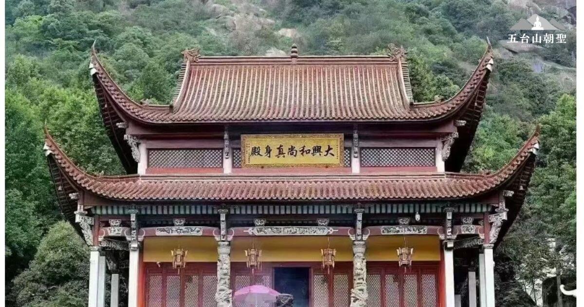 這張圖片展示了大興肉身殿（Daxing Flesh Body Hall）壯麗的建築外觀，這座殿堂坐落在山間，四周環繞著翠綠的山景。建築的屋頂呈現出典型的中國古代寺廟風格，紅色和金色的裝飾增添了神聖與莊嚴的氛圍。殿堂上方的匾額寫著「五高和照大」，這是對寺廟所代表的宗教意義的讚譽。這座建築的設計充滿了中國傳統文化的象徵，是許多朝聖者與遊客前來參拜與瞻仰的地點。