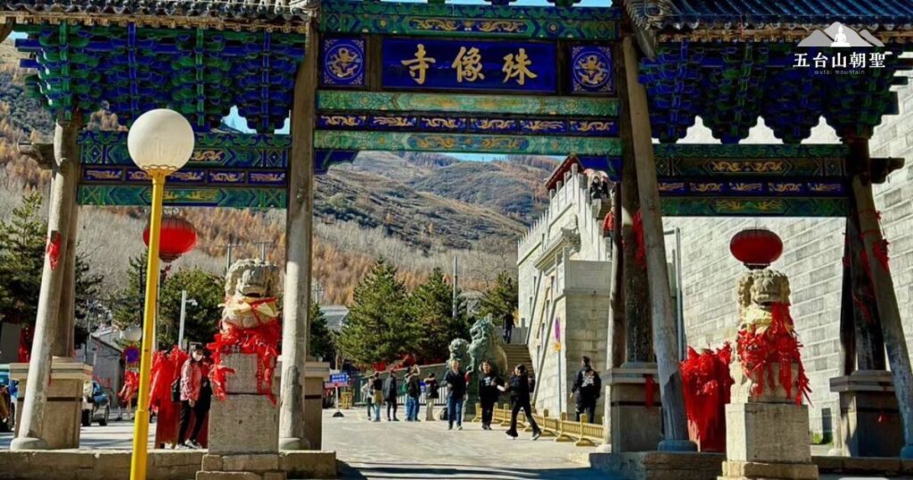 五台山著名的殊像寺入口牌樓。，這座藍色琉璃瓦的古典牌樓上寫著「殊像寺」，象徵著佛教聖地的尊貴與莊嚴。門口的石獅子綁著紅色祈願帶，展現朝聖文化與信仰氛圍。背景可見遠山環繞，象徵五台山作為佛教四大名山之一的殊勝地位。畫面中，遊客與信徒穿越寺門，象徵著五台山作為朝聖地的歷史與現代融合，是旅遊與信仰的交匯之地。