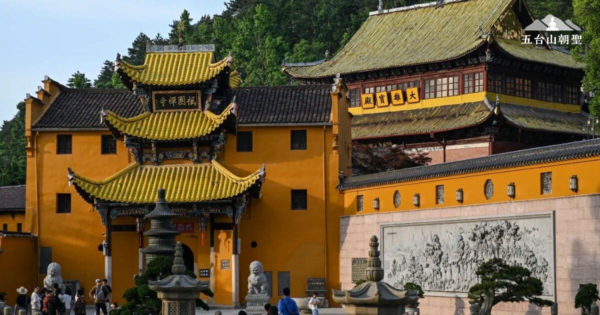 這張圖片展示的是位於山間的戈音寺，寺廟的建築融合了中國傳統的黃金瓦屋頂與橙色牆面，給人一種莊嚴、安詳的感覺。高聳的屋頂、精緻的雕刻和古老的風格讓這座寺廟更顯神聖。前景中是石獅和精緻的雕刻牆，這些設計向訪客展示了深厚的文化背景。四周的綠樹和晴朗的天空，為這座寺廟增添了自然的和諧與靜謐氛圍。