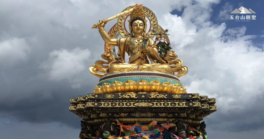 這張圖片展示了**五台山(Wutai Mountain)的文殊菩薩(Manjushri Bodhisattva)**金色雕像,菩薩端坐於精美的蓮花座上,右手高舉智慧之劍,象徵斬斷無明與煩惱,左手持蓮花,展現清淨與智慧的象徵。雕像金光閃耀,細節雕刻精緻,底座裝飾華麗,搭配周圍色彩繽紛的經幡,增添了濃厚的佛教氛圍。背景中雲層翻湧,象徵五台山的靈性與神秘感,這座雕像吸引無數朝聖者前來禮拜,祈求智慧與加持。這張圖片適合用於介紹五台山的宗教文化與文殊菩薩的象徵意義。
