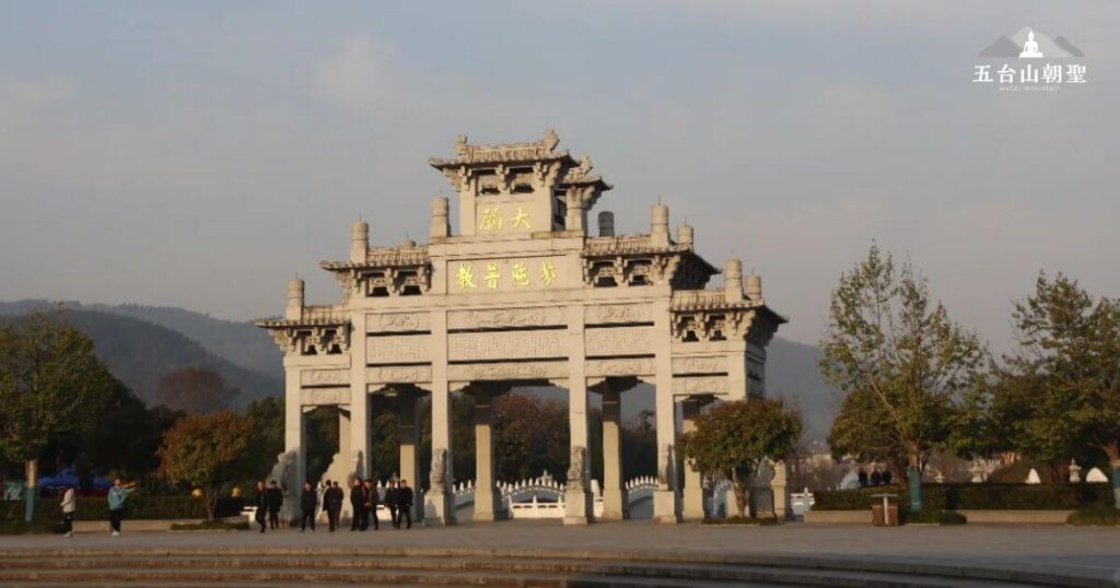 這張圖片展示的是洪源大殿的正門，這座建築的典雅氣勢顯示了傳統的中國古建築風格。大殿的拱形門樓雕刻精美，四周的環境綠樹成蔭，空氣清新。門上方寫著「顯大教音遷學」的金色大字，透露出這座殿堂的文化底蘊和歷史傳承。背景的山脈和悠閒的人群，使這個場所充滿了和諧與寧靜的氛圍。