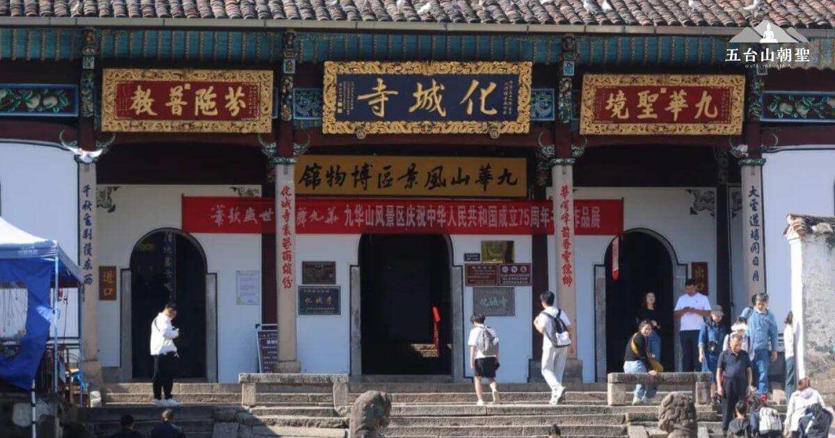 這張圖片展示的是花城寺的正門，寺廟的建築風格典雅，展現了傳統中國的寺廟特色。門上掛著精美的匾額，分別書寫著「教誨隨光」和「境聖華九」，為寺廟增添了莊嚴的氛圍。圖片中可以看到遊客在參觀這座寺廟，並且有一些紅色的橫幅標誌著重要的紀念活動，如慶祝中華人民共和國建國75週年等。寺廟的門前石獅和階梯使整個場景更加充滿古老的文化韻味。