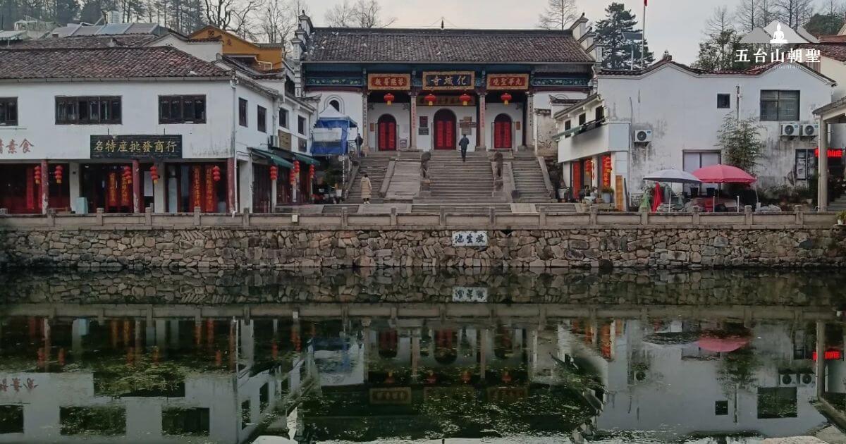 這張圖片展示了九華街（Jiuhua Street）上古老建築的景觀，反射在水面上的景象更是為這一切增添了詩意。石階通向大廳，兩側的商店則展示著中國傳統的街市風貌，紅色的燈籠裝飾著街道，為這座歷史悠久的街區增添了一抹節日氣息。這裡的景色不僅吸引著遊客，也讓人感受到濃厚的文化底蘊。