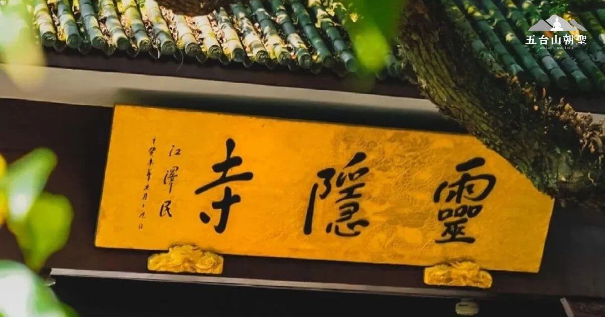 這張圖片展示了靈隱寺（Lingyin Temple）的大門匾額。匾額上用金色的字寫著「靈隱」，這座寺廟位於浙江省杭州市，是中國佛教禪宗的著名寺廟之一。背景中可以看到古老的瓦片屋頂，並且匾額的邊緣被綠意盎然的樹枝環繞，營造出一種寧靜、神聖的氛圍。這個標誌性的建築吸引了無數朝聖者與遊客前來參拜與欣賞。