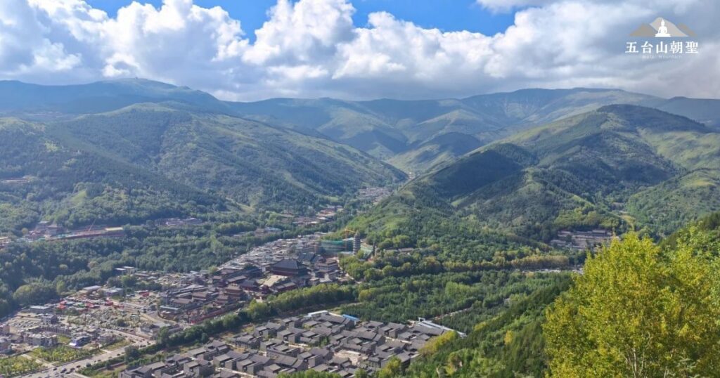 這張圖片展示了五台山的壯麗自然景觀，山脈起伏，綠意盎然。五台山是佛教朝聖的聖地，擁有3061米的海拔高度，這使得它比台灣的玉山還要高。畫面中可以看到五台山的山脈和遠處的建築群，這些建築群大多與佛教文化有關，是信徒來此朝聖與靈性修行的重要場所。