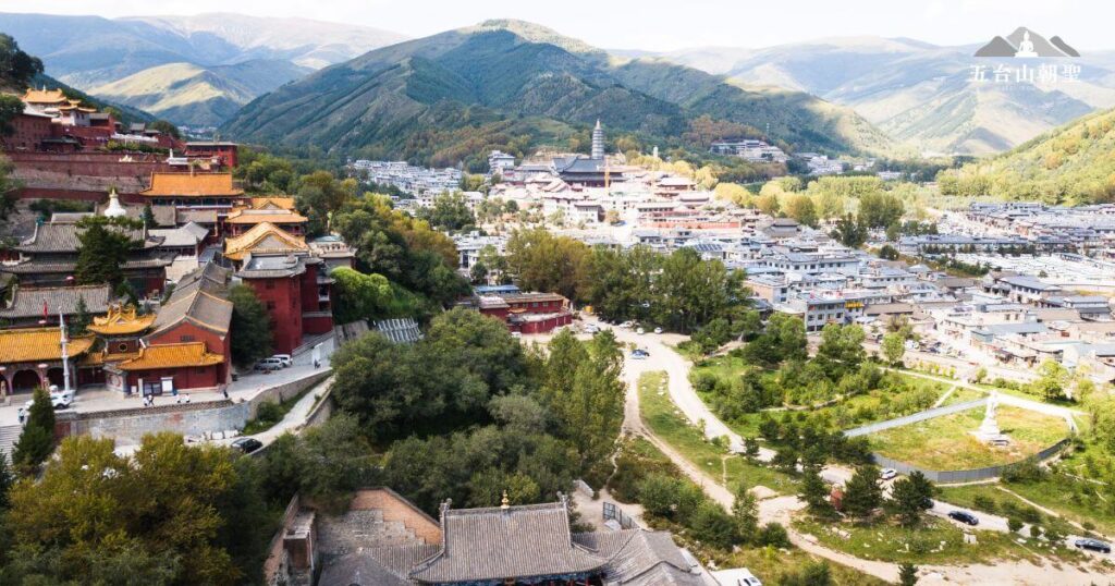 這張圖片展示了五台山（Wutai Mountain）的壯麗全景，包含傳統的佛教寺廟建築群、綠意盎然的山脈與現代城鎮交錯的景色。左側可見層層堆疊的紅色與金色屋頂的寺廟建築，展現濃厚的宗教氛圍，而中央遠處的塔院寺白塔成為視覺焦點，象徵五台山作為文殊菩薩道場的重要地位。遠方綿延起伏的山脈環抱整座聖地，使五台山成為朝聖與修行的絕佳之地。