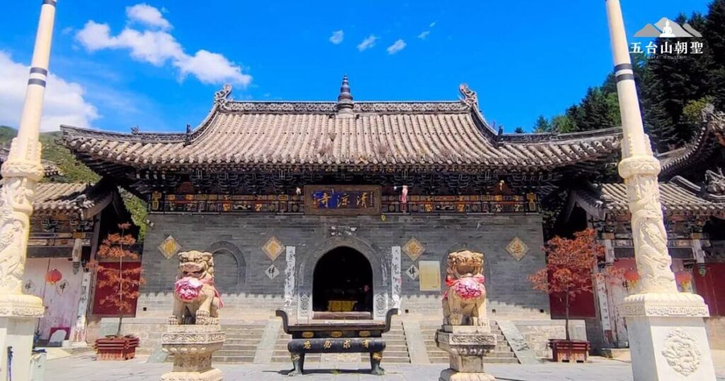 這張圖片展示了五台山清涼寺的正門，灰色磚牆與精美的木雕裝飾突顯了古寺的歷史感。門前兩座石獅系上紅色綢帶，象徵著吉祥與守護，而香爐則顯示出信眾的虔誠供奉。藍天下的清涼寺顯得格外莊嚴肅穆，兩側白色石柱雕刻精美，增添了佛教聖地的神聖氛圍。這座寺廟是朝聖者修行與靜心的理想之地，讓旅人感受佛教文化的深厚底蘊。