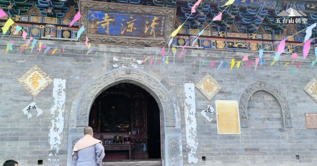 這張照片展示了五台山清涼寺的正門，古樸的灰磚牆面與雕花石門框，展現了歷史的滄桑感。門額上方的「清涼寺」匾額金字熠熠生輝，與色彩豐富的木雕裝飾形成對比，增添莊嚴氛圍。門口一名僧侶靜靜佇立，展現朝聖者虔誠的信仰。上方懸掛著五彩經幡，象徵佛法的無邊庇佑，使這座寺廟更顯靈性與寧靜。