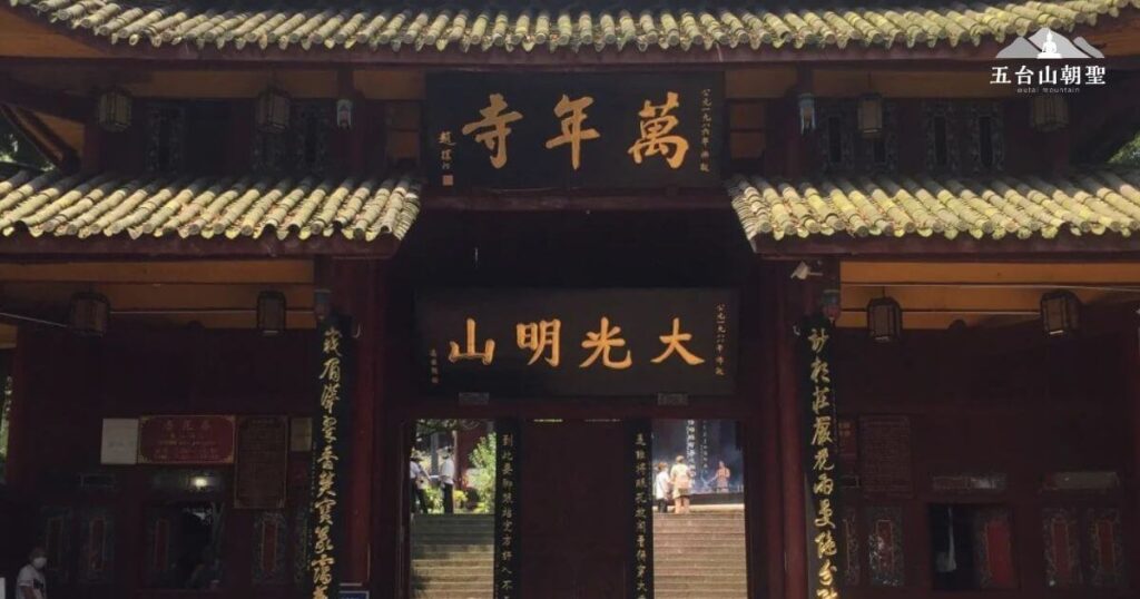 這張圖片展示的是位於峨眉山的萬年寺。萬年寺是中國四川省峨眉山的一個重要佛教寺廟，擁有深厚的宗教背景和歷史文化。寺廟的大門上方的匾額上寫著「山明光大」，這意味著佛法光明廣大，普照眾生。萬年寺以其悠久的歷史、壯麗的建築以及其所承載的佛教文化而聞名，是許多信徒朝聖的目的地。這裡的建築風格典雅，與周圍的自然景色融合，營造出寧靜而神聖的氛圍。