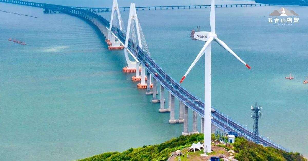 這張圖片展示了位於中國舟山群島的舟山跨海大橋（Zhoushan cross-sea bridge）。這座橋梁是中國的重要交通設施，跨越海域連接舟山與浙江本土。照片中可以看到橋樑與一座大型風力發電機共同構成的現代化景觀，背景中的海面與自然景色融為一體，顯示出人類與自然的和諧共存。